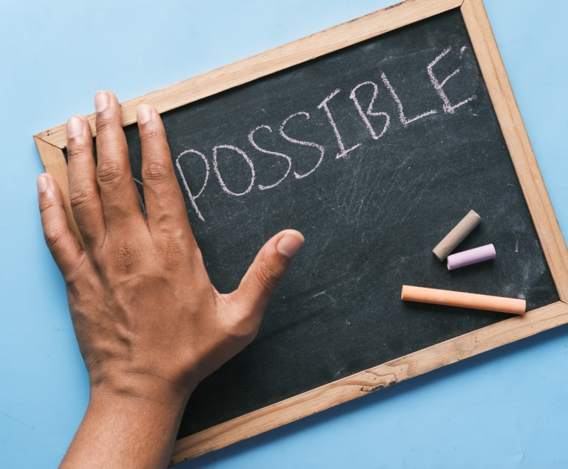A left hand is rested on a small blackboard on a surface. On the board is written 'possible' and the hand appears to be covering up the letters 'Im'