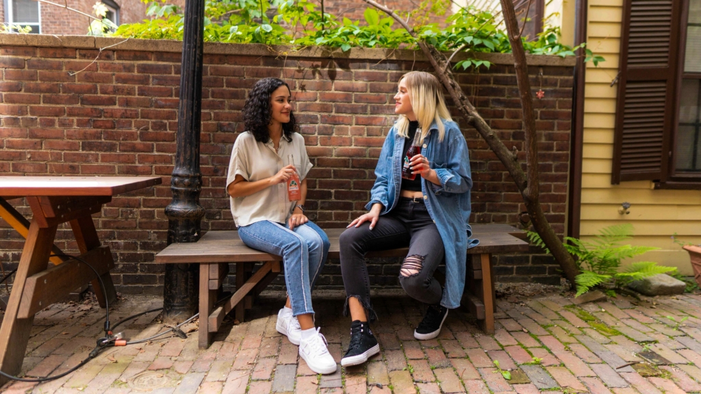 Two young women sit beside each other on a bench in a paved courtyard have a coffee and a conversation