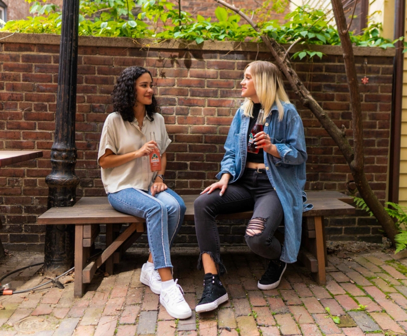 Two young women sit beside each other on a bench in a paved courtyard have a coffee and a conversation